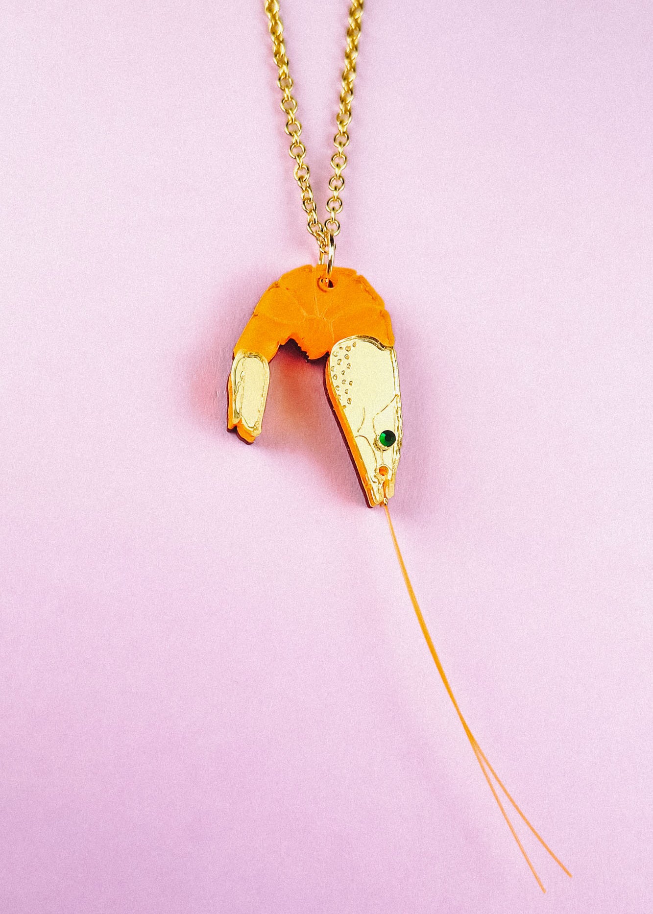 A whimsical Shrimp Pendant Necklace: this gold necklace features a playful orange shrimp pendant with a green gem eye and thin gold whiskers, beautifully displayed on a pastel pink background.