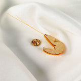 A Shrimp Pin accessory with orange accents and a gold pin is displayed on a soft, white, textured fabric background, adding a playful wardrobe charm to any outfit.