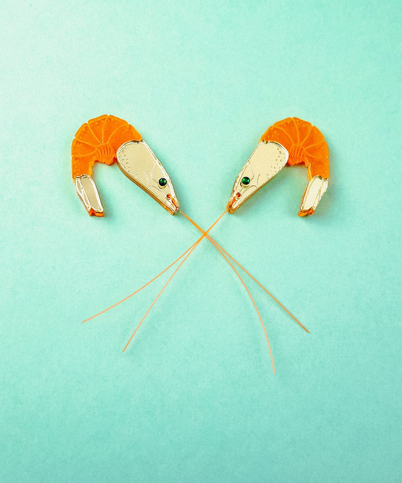 Two neon orange Shrimp Stud earrings, shaped like shrimp with long antennae crossing in the center, are positioned facing each other on a turquoise background. Subtle emerald rhinestones add a touch of sparkle to their playful design.