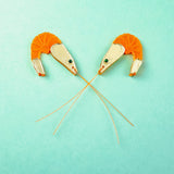 Two neon orange Shrimp Stud earrings, shaped like shrimp with long antennae crossing in the center, are positioned facing each other on a turquoise background. Subtle emerald rhinestones add a touch of sparkle to their playful design.