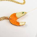 A playful Shrimp Pendant Necklace with an orange body and gold claw details rests on a white surface, showcasing its whimsical design.