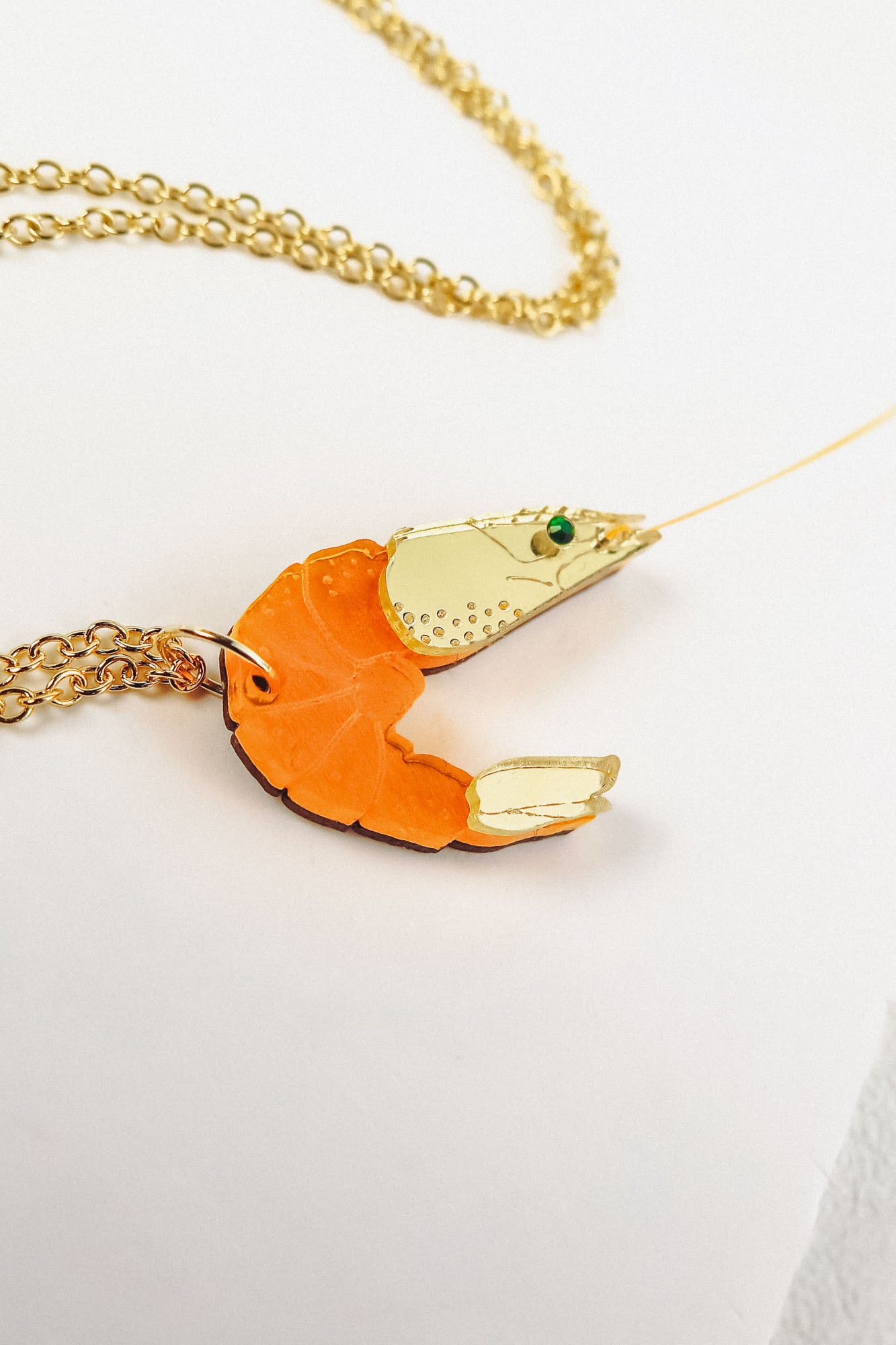 A playful Shrimp Pendant Necklace with an orange body and gold claw details rests on a white surface, showcasing its whimsical design.