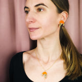 A woman with light skin and straight brown hair in a low ponytail wears a playful black top, a whimsical Shrimp Pendant Necklace, and matching gold and orange earrings, standing in front of a mauve curtain.