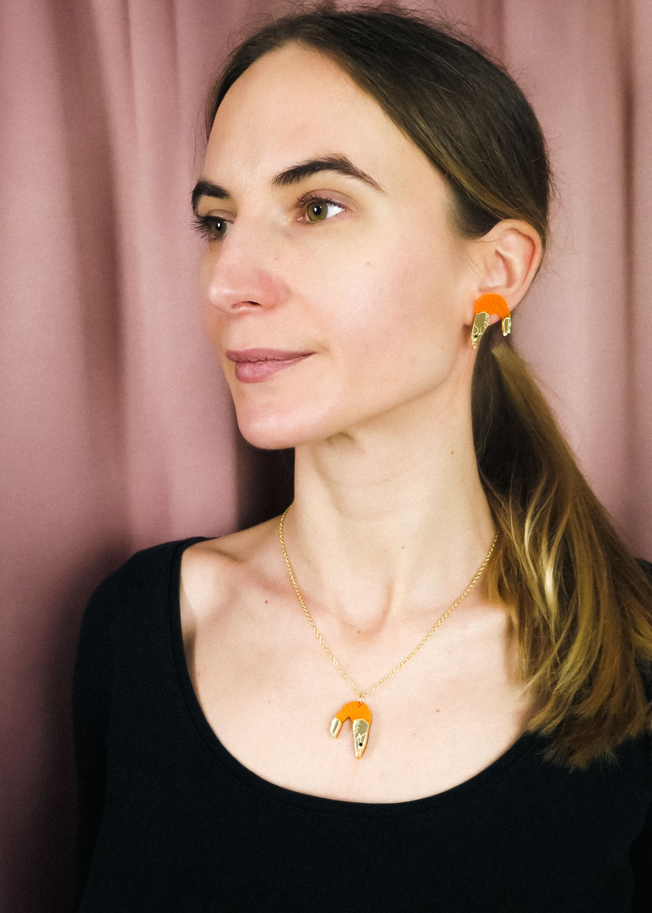 A woman with light skin and straight brown hair in a low ponytail wears a playful black top, a whimsical Shrimp Pendant Necklace, and matching gold and orange earrings, standing in front of a mauve curtain.