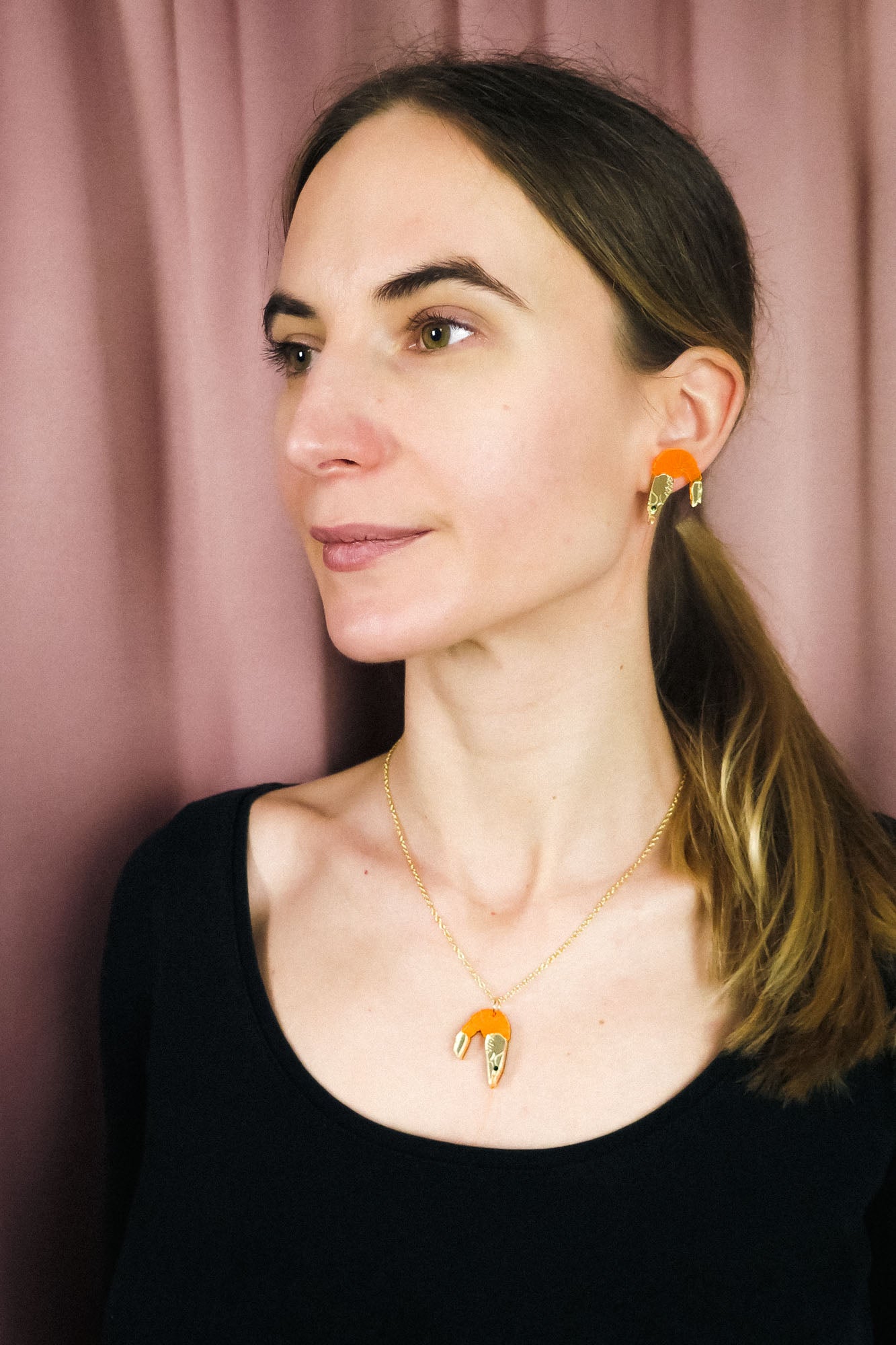 A woman with light skin and straight brown hair in a low ponytail wears a playful black top, a whimsical Shrimp Pendant Necklace, and matching gold and orange earrings, standing in front of a mauve curtain.