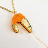 A playful Shrimp Pendant Necklace with a gold chain features an orange shrimp pendant adorned with gold accents and a green gem eye, creating a whimsical touch against a white background.