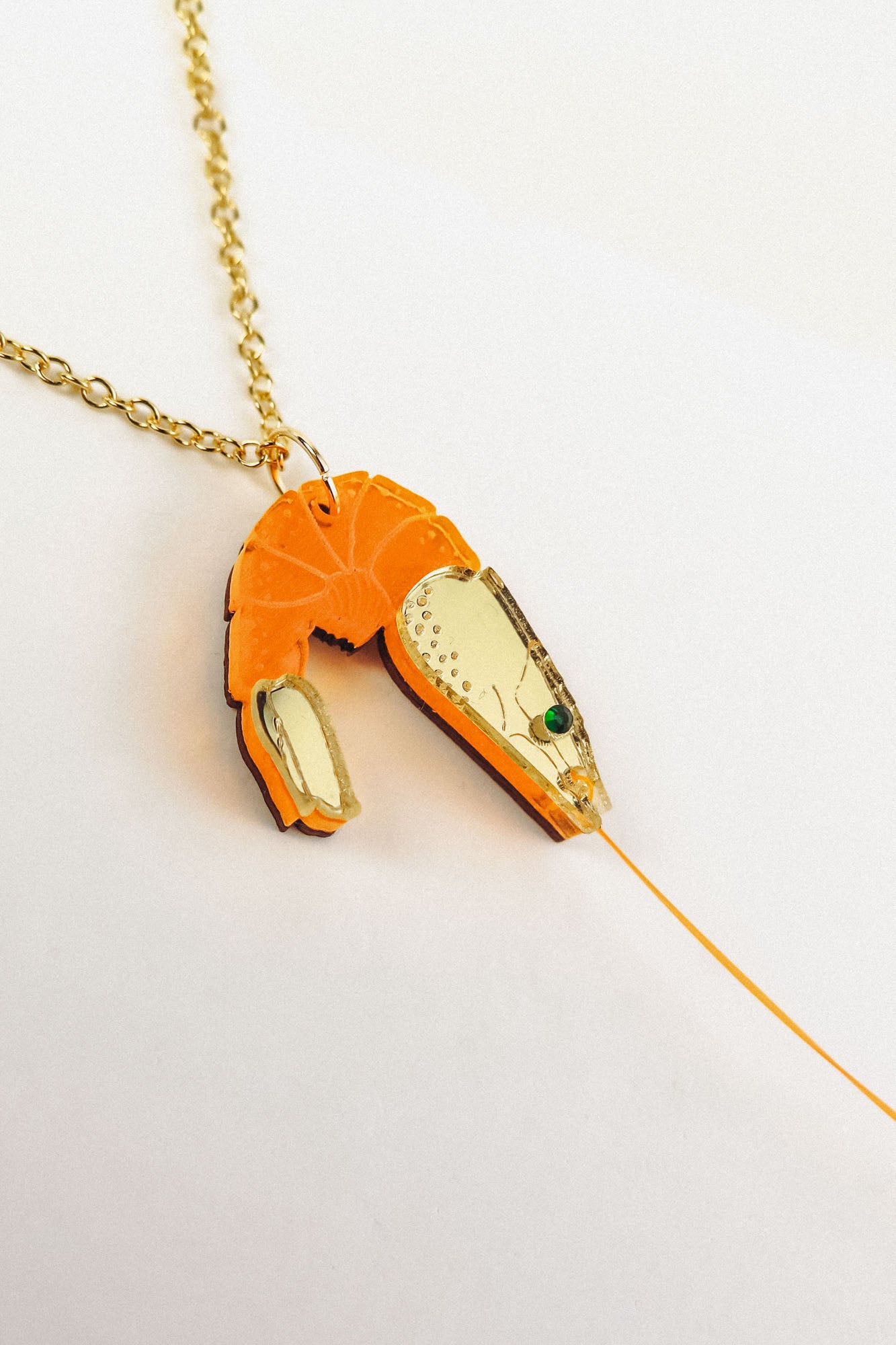 A playful Shrimp Pendant Necklace with a gold chain features an orange shrimp pendant adorned with gold accents and a green gem eye, creating a whimsical touch against a white background.