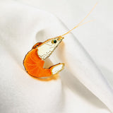 A decorative orange and clear glass shrimp figurine, perfect as an accessory or unique wardrobe charm, rests on white, softly folded fabric, showcasing fine details on its body and long antennae extending outward.