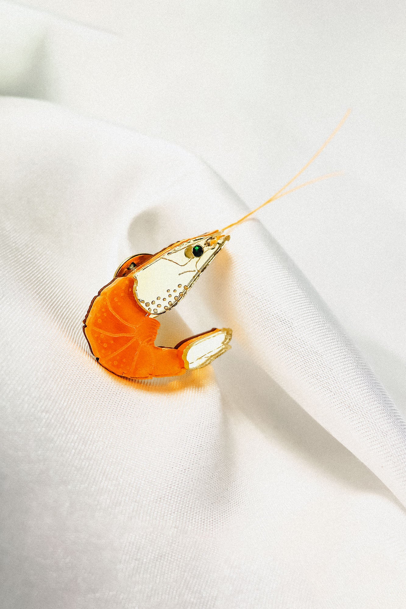 A decorative orange and clear glass shrimp figurine, perfect as an accessory or unique wardrobe charm, rests on white, softly folded fabric, showcasing fine details on its body and long antennae extending outward.
