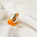 A shrimp-shaped brooch accessory with a shiny silver and orange design, small green gemstone detail, and two thin antennae—this Shrimp Pin brings playful wardrobe charm as it rests on white satin fabric.