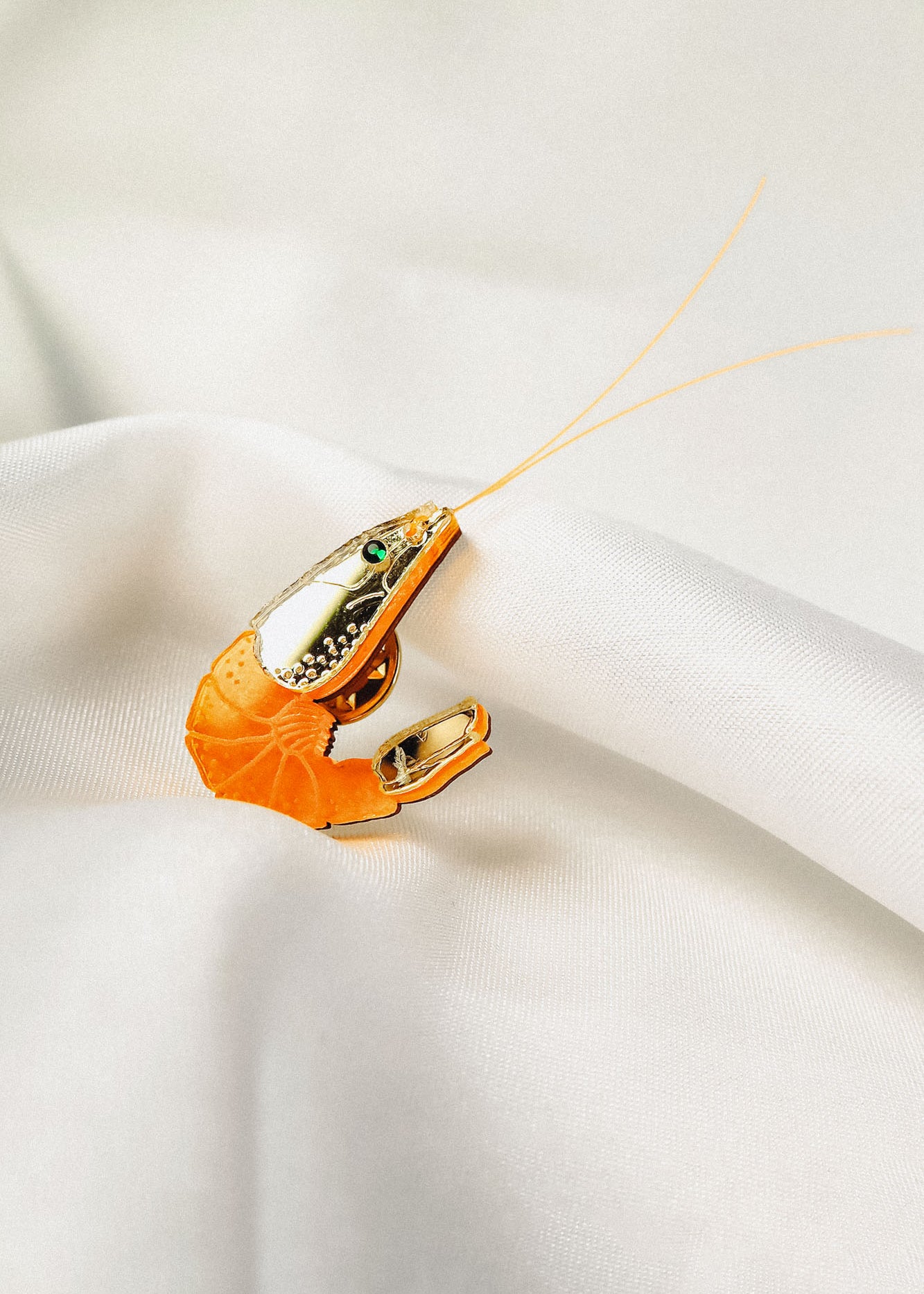 A shrimp-shaped brooch accessory with a shiny silver and orange design, small green gemstone detail, and two thin antennae—this Shrimp Pin brings playful wardrobe charm as it rests on white satin fabric.