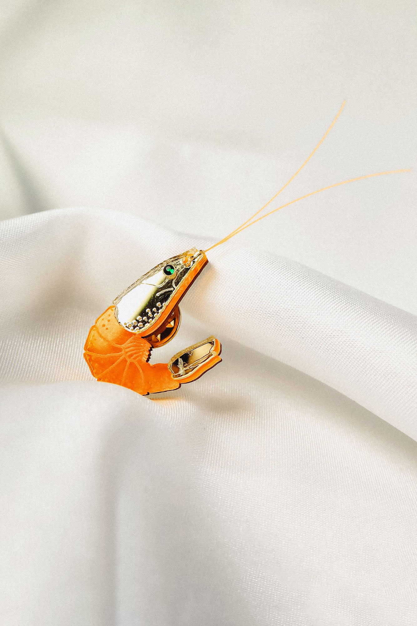 A shrimp-shaped brooch accessory with a shiny silver and orange design, small green gemstone detail, and two thin antennae—this Shrimp Pin brings playful wardrobe charm as it rests on white satin fabric.