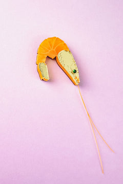 A Shrimp Pin with orange, gold, and green details is placed on a light purple background. Long thin strings extend from the brooch, mimicking shrimp antennae—making it a playful accessory and unique wardrobe charm.