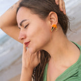 A woman with long brown hair and a green shirt stands on a beach with her eyes closed, smiling gently, and one arm raised over her head, showing off Shrimp Stud earrings sparkling with emerald rhinestones.
