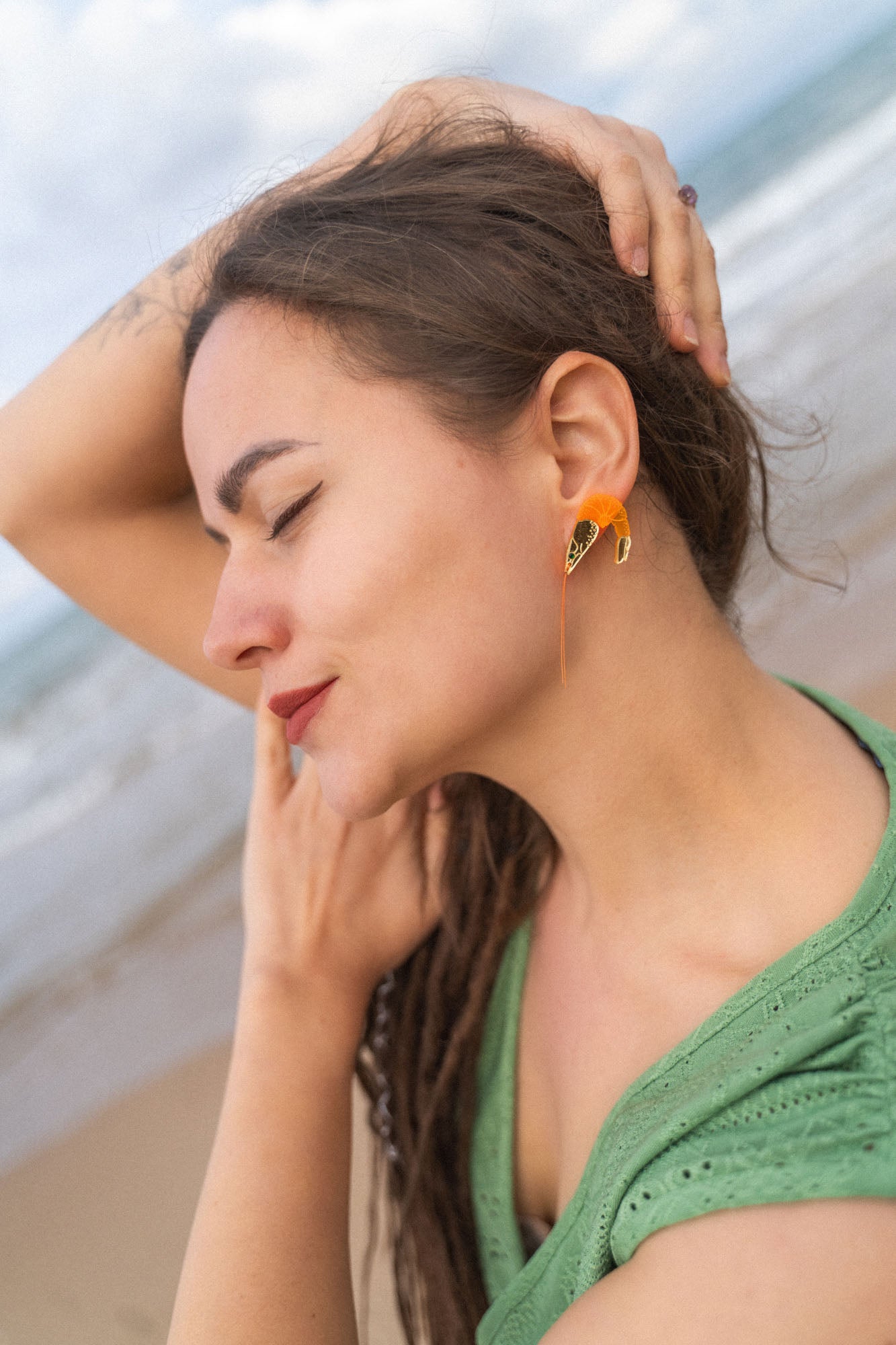 A woman with long brown hair and a green shirt stands on a beach with her eyes closed, smiling gently, and one arm raised over her head, showing off Shrimp Stud earrings sparkling with emerald rhinestones.