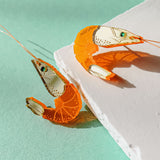 Two neon orange Shrimp Stud earrings, possibly adorned with emerald rhinestones, rest on a textured white surface against a mint green background, casting distinct shadows.