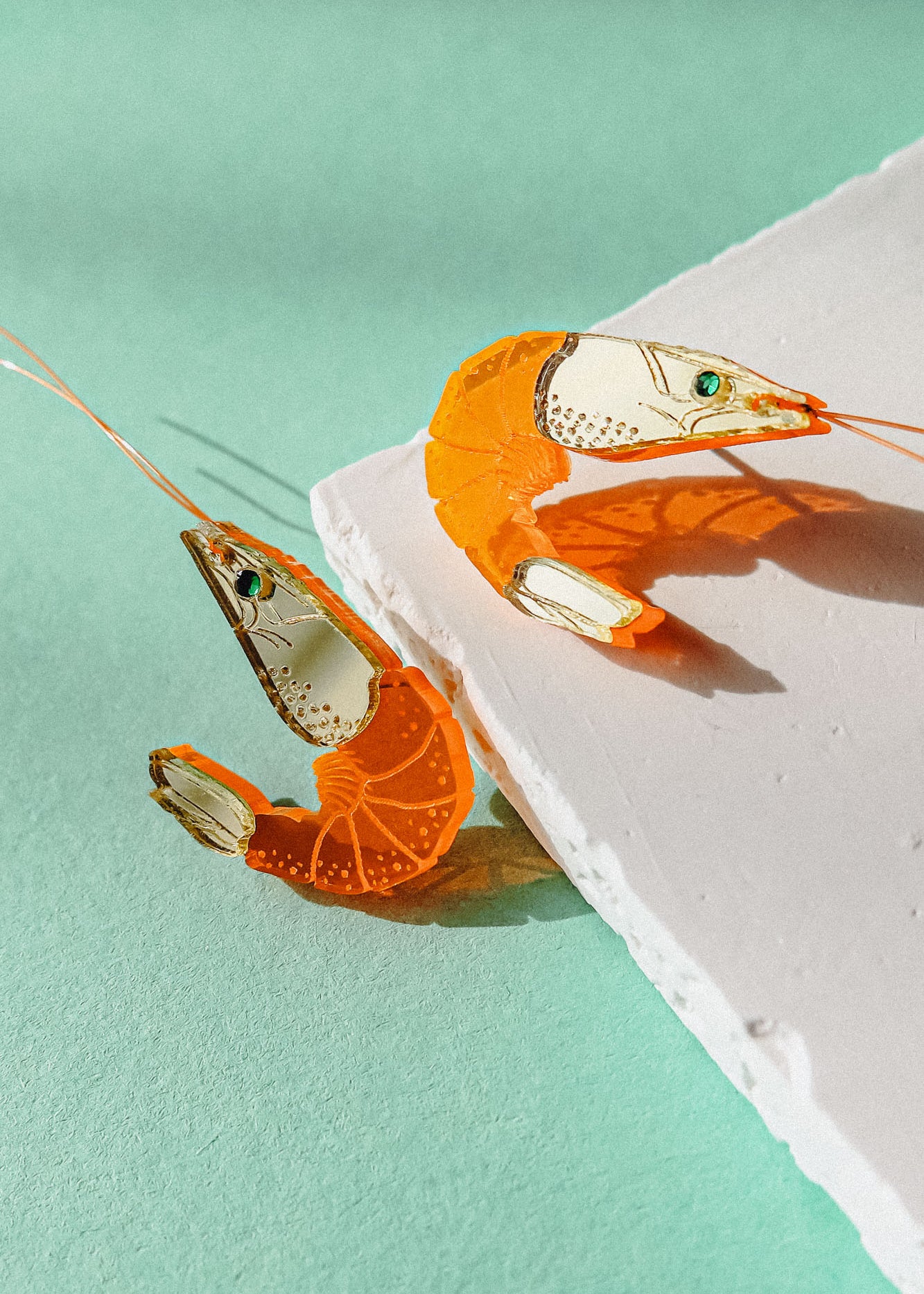 Two neon orange Shrimp Stud earrings, possibly adorned with emerald rhinestones, rest on a textured white surface against a mint green background, casting distinct shadows.