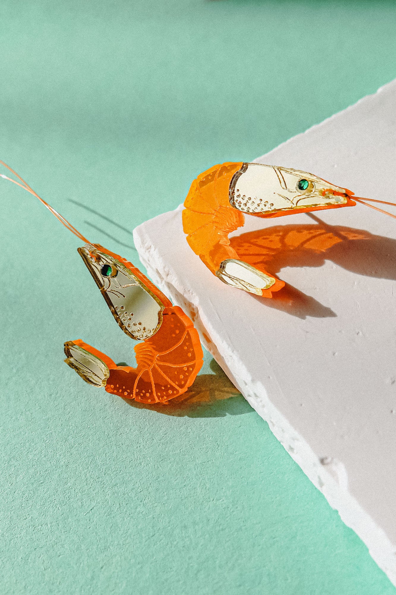 Two neon orange Shrimp Stud earrings, possibly adorned with emerald rhinestones, rest on a textured white surface against a mint green background, casting distinct shadows.