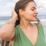 A woman with long brown hair in a green dress stands on a beach, eyes closed and smiling slightly, with one hand touching her hair. She wears neon orange Shrimp Stud earrings and has a tattoo on her arm. The ocean is in the background.