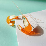 A peeled shrimp with its shell partially removed lies on a white surface against a mint green background, casting a shadow in bright sunlight—its curves reminiscent of elegant Shrimp Stud earrings.