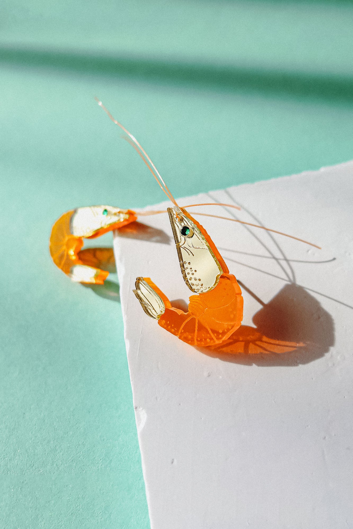 A peeled shrimp with its shell partially removed lies on a white surface against a mint green background, casting a shadow in bright sunlight—its curves reminiscent of elegant Shrimp Stud earrings.