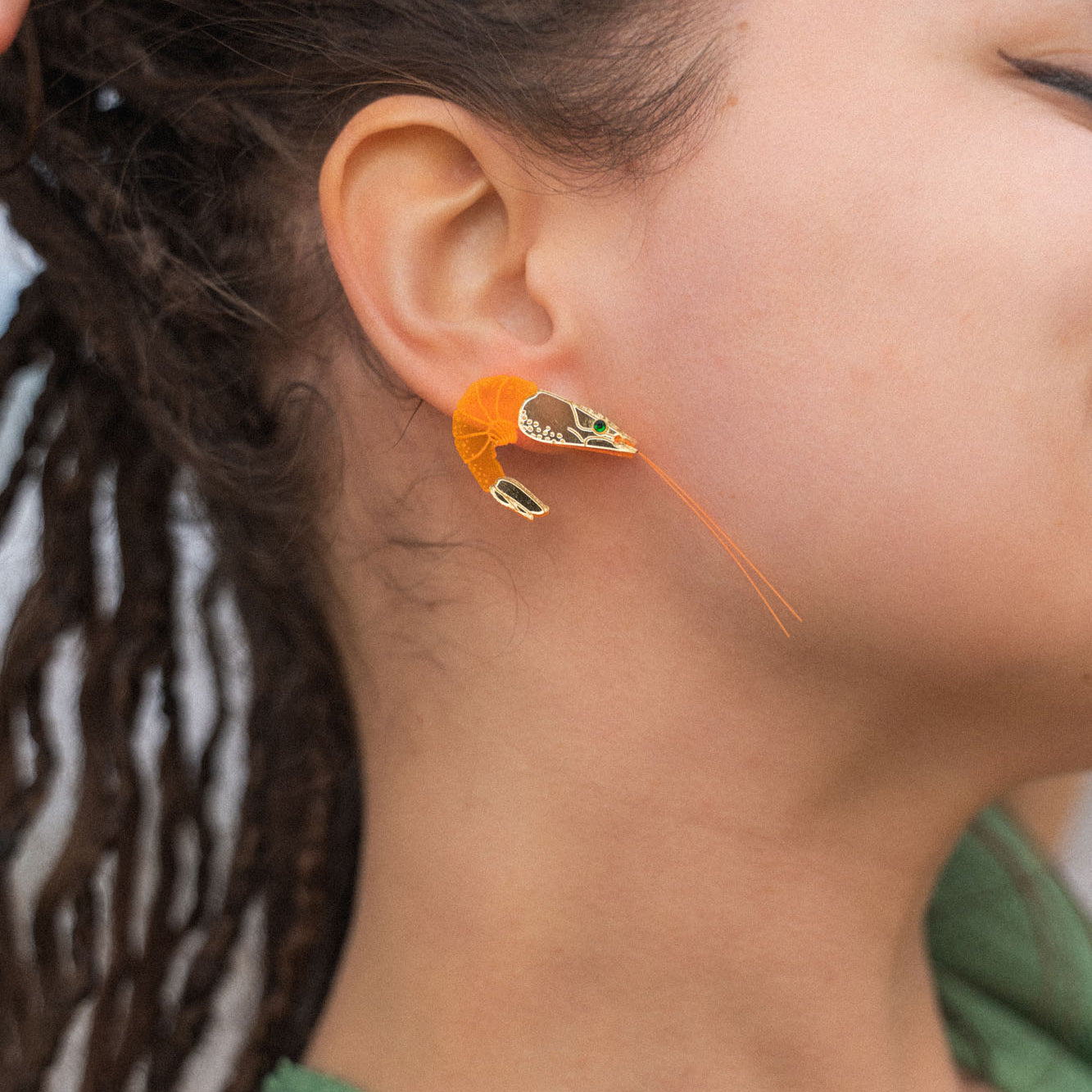 A person with brown skin and dreadlocks wears a detailed, neon orange Shrimp Stud earring adorned with emerald rhinestones. Their hand rests on their head, and they are dressed in a green top.