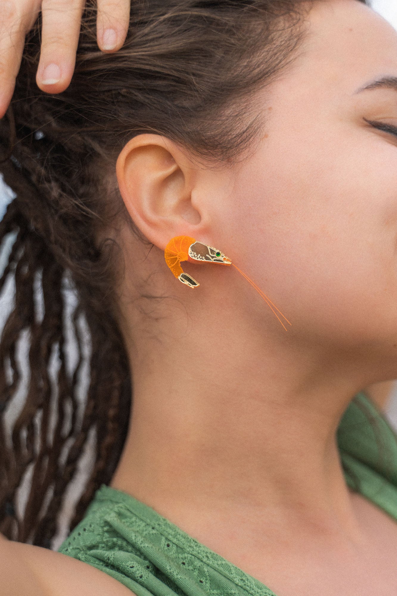 A person with brown skin and dreadlocks wears a detailed, neon orange Shrimp Stud earring adorned with emerald rhinestones. Their hand rests on their head, and they are dressed in a green top.