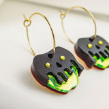 Limited edition Poisoned Apple Earrings with gold hoops and dangling black and neon green charms. Featuring a Halloween design with stylized dripping details and yellow dot accents, these earrings rest elegantly on a white surface.