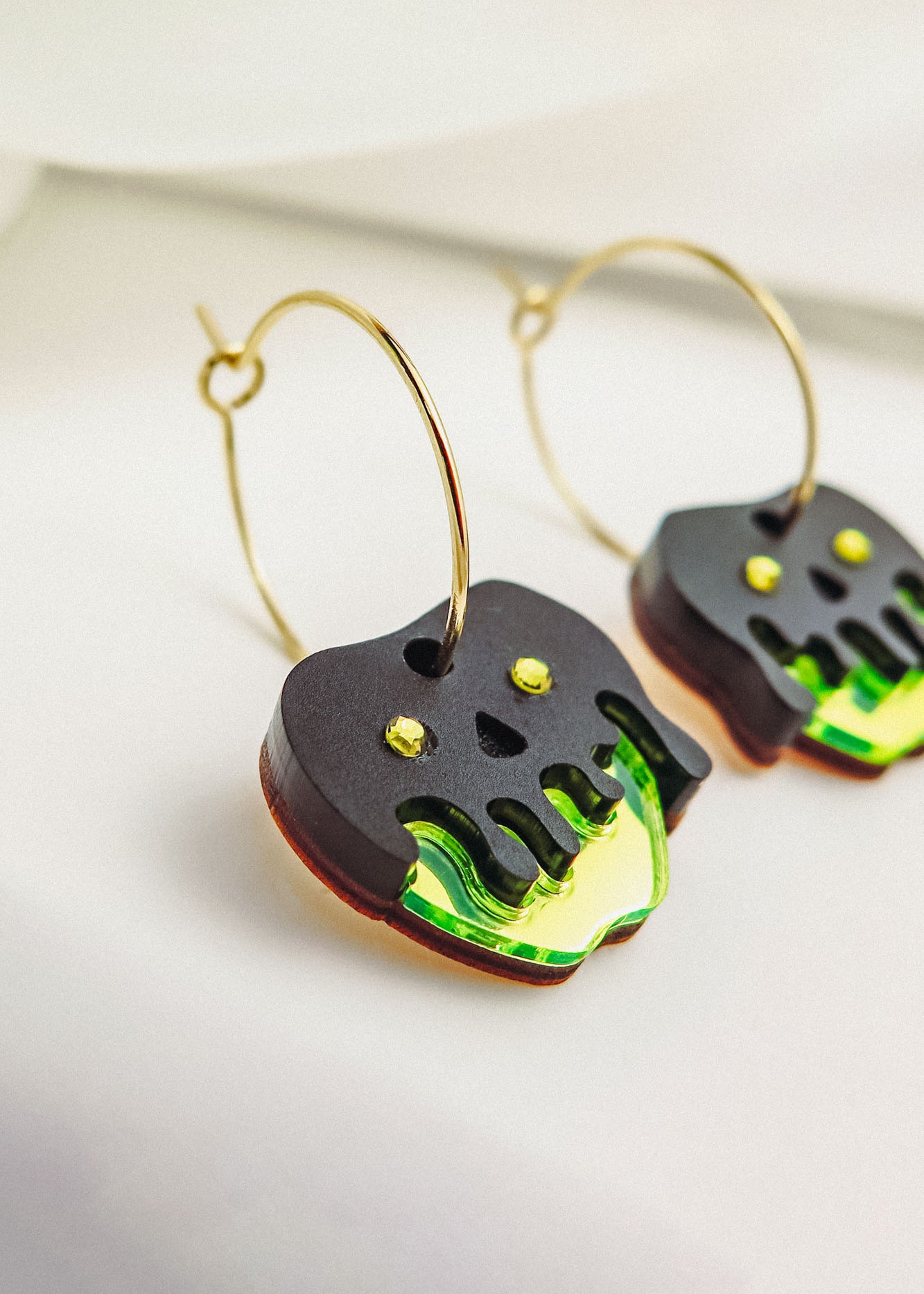 Limited edition Poisoned Apple Earrings with gold hoops and dangling black and neon green charms. Featuring a Halloween design with stylized dripping details and yellow dot accents, these earrings rest elegantly on a white surface.