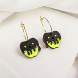 A pair of limited edition gold hoop earrings featuring black apple-shaped charms with bright green dripping accents, inspired by a Poisoned Apple Earrings Halloween design, displayed on a white fabric background.