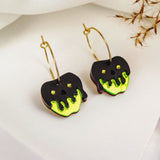 A pair of Poisoned Apple Earrings shaped like apples, with black and neon green dripping designs resembling poison, hang from gold hoops on a white surface with dried grass in the background—a limited edition Halloween design.