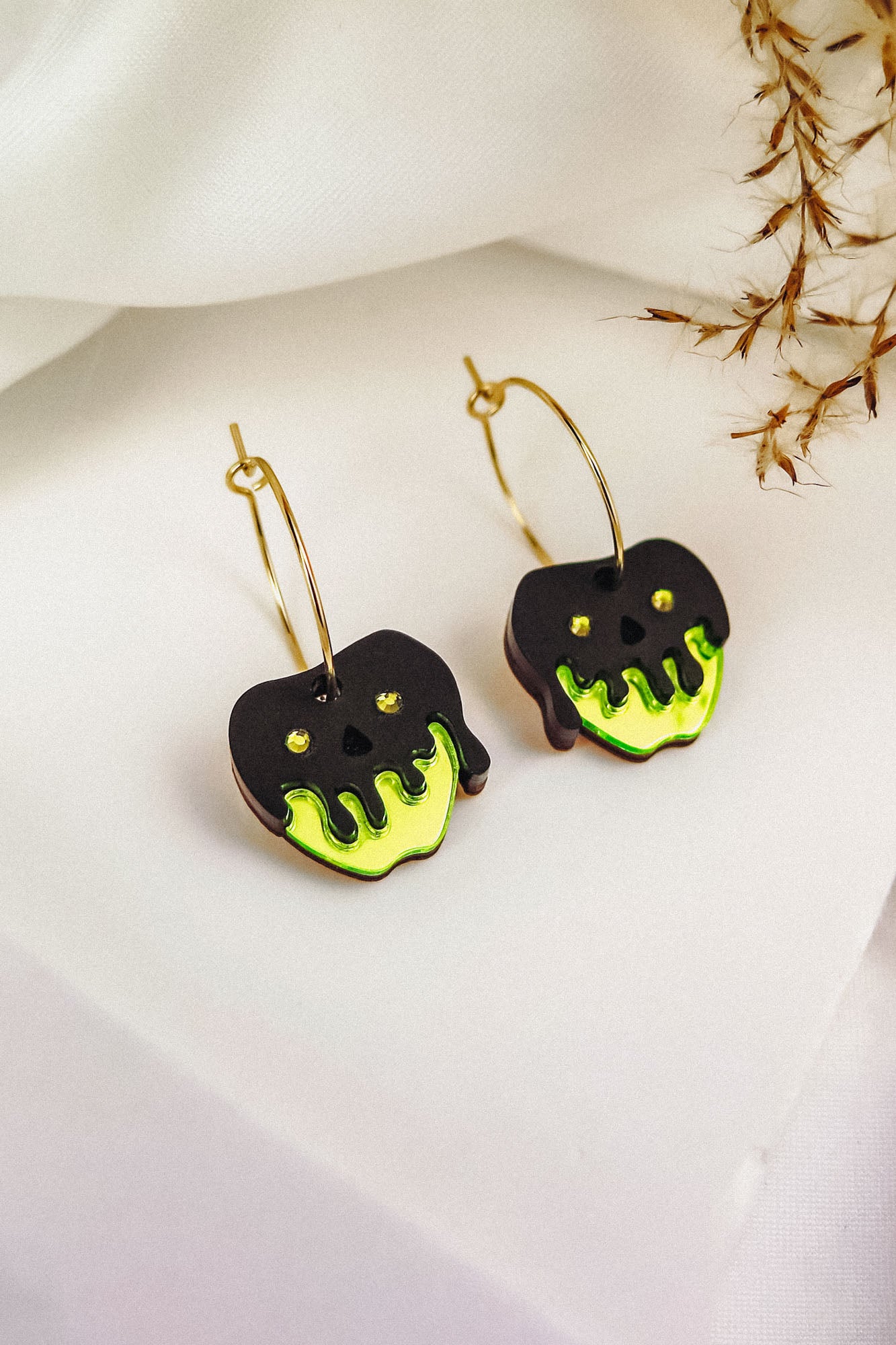 A pair of Poisoned Apple Earrings shaped like apples, with black and neon green dripping designs resembling poison, hang from gold hoops on a white surface with dried grass in the background—a limited edition Halloween design.