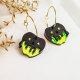 A pair of Poisoned Apple Earrings with gold hoops and black skull-shaped charms, decorated with bright green, dripping accents and small yellow dots in a Halloween design, are displayed on a white surface near dried plants.