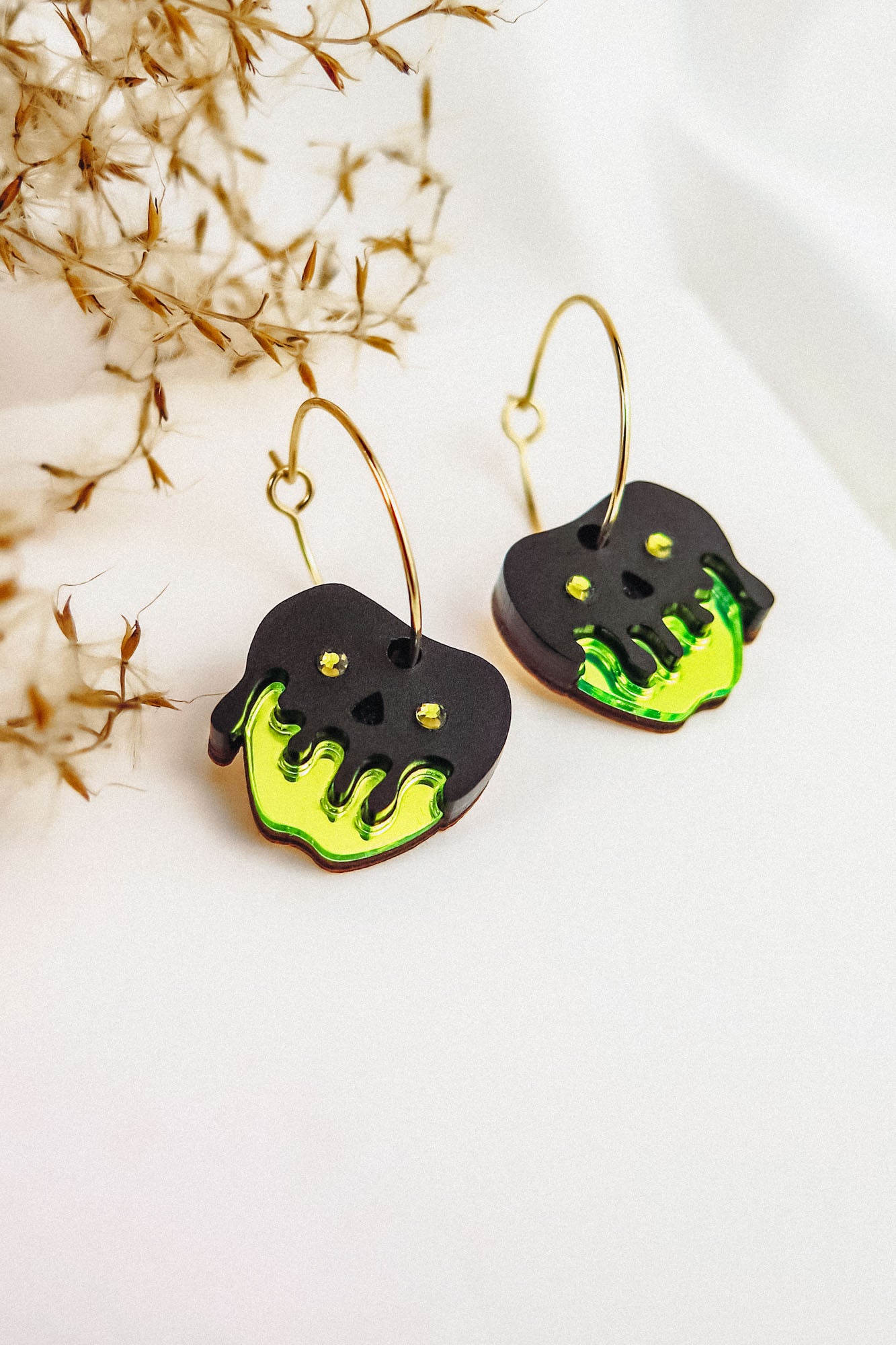 A pair of Poisoned Apple Earrings with gold hoops and black skull-shaped charms, decorated with bright green, dripping accents and small yellow dots in a Halloween design, are displayed on a white surface near dried plants.