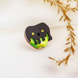 A Poisoned Apple Badge pin shaped like a skull with black and green melting details, featuring yellow eyes and nose holes. Perfect for Halloween lovers, its backdrop is softly blurred with dried grass on the right. Limited stock available!.