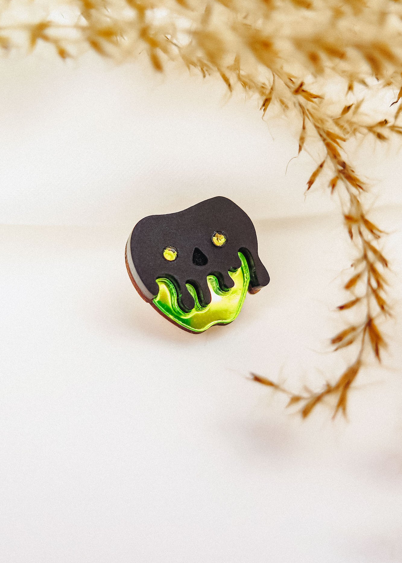 A Poisoned Apple Badge pin shaped like a skull with black and green melting details, featuring yellow eyes and nose holes. Perfect for Halloween lovers, its backdrop is softly blurred with dried grass on the right. Limited stock available!.