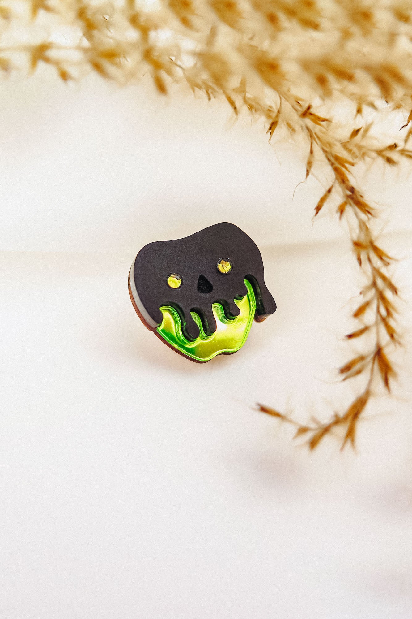 A Poisoned Apple Badge pin shaped like a skull with black and green melting details, featuring yellow eyes and nose holes. Perfect for Halloween lovers, its backdrop is softly blurred with dried grass on the right. Limited stock available!.