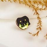 A small Poisoned Apple Badge shaped like a black skull with glowing green accents rests on white fabric, surrounded by delicate, dried plant stems—perfect for Halloween lovers and available in limited stock.