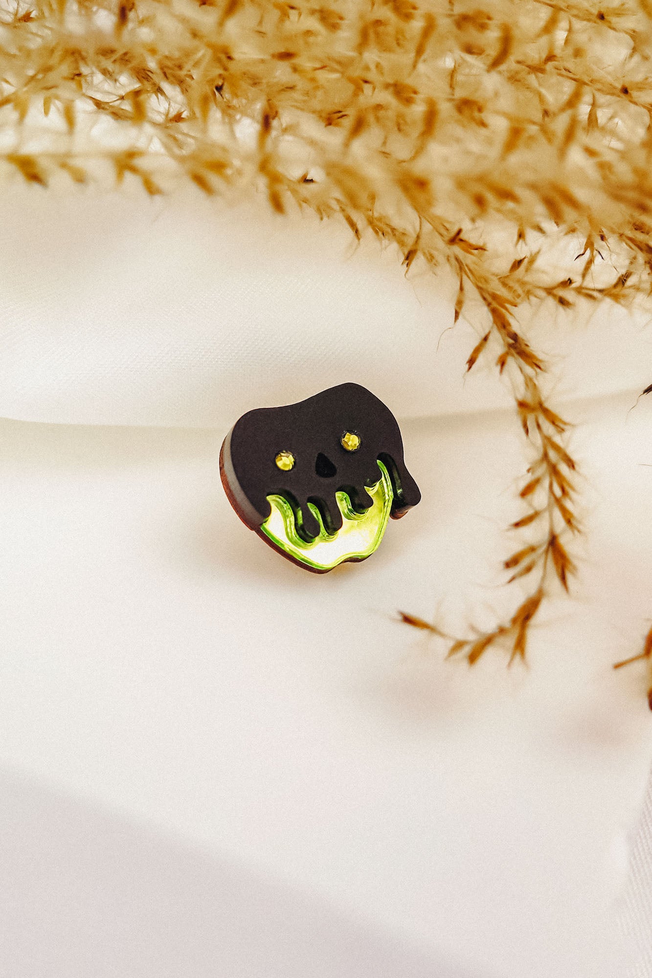 A small Poisoned Apple Badge shaped like a black skull with glowing green accents rests on white fabric, surrounded by delicate, dried plant stems—perfect for Halloween lovers and available in limited stock.