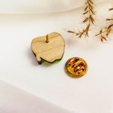 A wooden enamel Poisoned Apple Badge pin, shown from the back with its gold butterfly clutch beside it, rests on a white surface with dried grass in the corner—perfect for Halloween lovers. Limited stock available!.