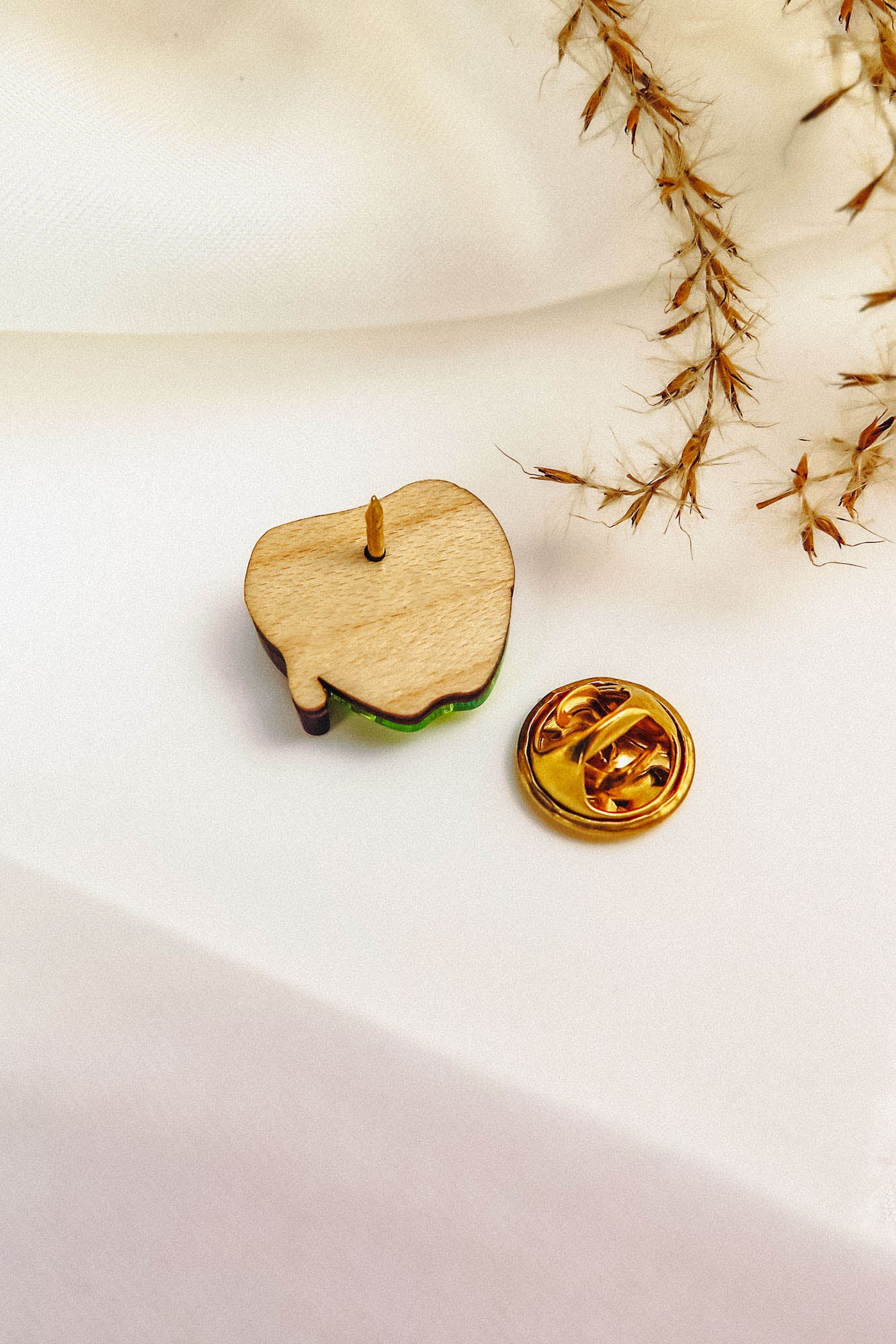 A wooden enamel Poisoned Apple Badge pin, shown from the back with its gold butterfly clutch beside it, rests on a white surface with dried grass in the corner—perfect for Halloween lovers. Limited stock available!.
