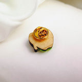 A wooden Poisoned Apple Badge pin with a gold clasp rests on a white fabric background. Perfect for Halloween lovers, its light brown surface features a green edge visible at the bottom. Limited stock available!.