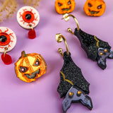 A set of Halloween-themed, hand-painted earrings on a purple surface, including pumpkin and eyeball earrings, a pumpkin brooch, and black bat-shaped earrings with glitter and purple accents. Dried plants are partially visible at the top.