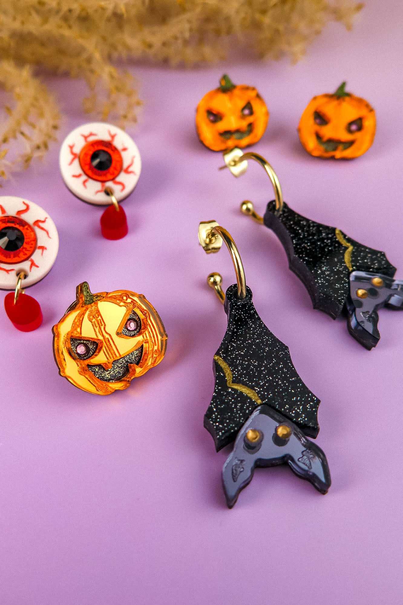 A set of Halloween-themed, hand-painted earrings on a purple surface, including pumpkin and eyeball earrings, a pumpkin brooch, and black bat-shaped earrings with glitter and purple accents. Dried plants are partially visible at the top.