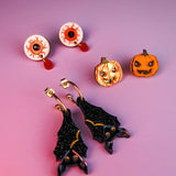 A set of festive Halloween earrings on a purple surface, including hand-painted earrings shaped like eyeballs, jack-o-lantern pumpkins, and sparkly black bats with gold accents—all allergy-free earrings for comfortable wear.