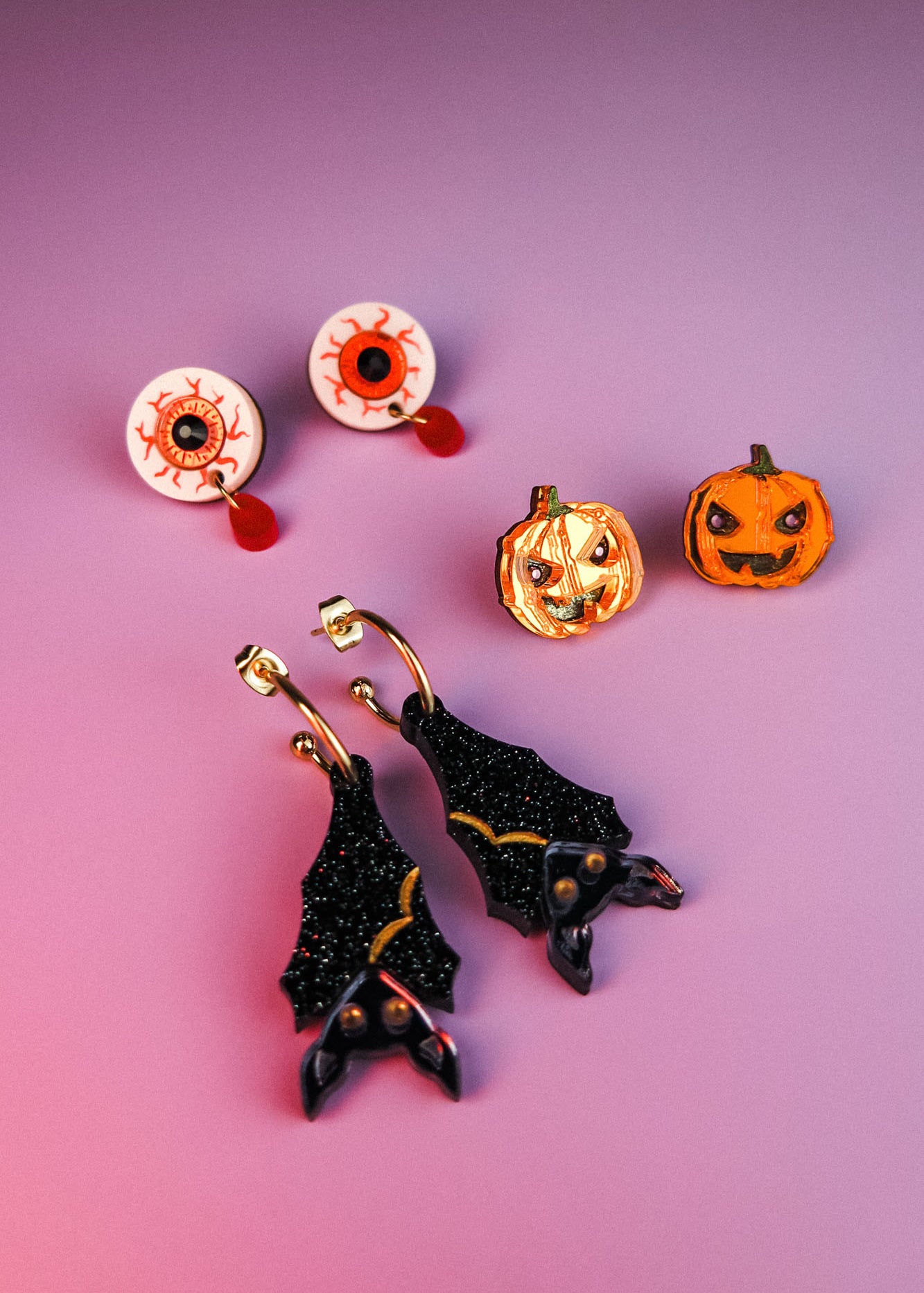 A set of festive Halloween earrings on a purple surface, including hand-painted earrings shaped like eyeballs, jack-o-lantern pumpkins, and sparkly black bats with gold accents—all allergy-free earrings for comfortable wear.