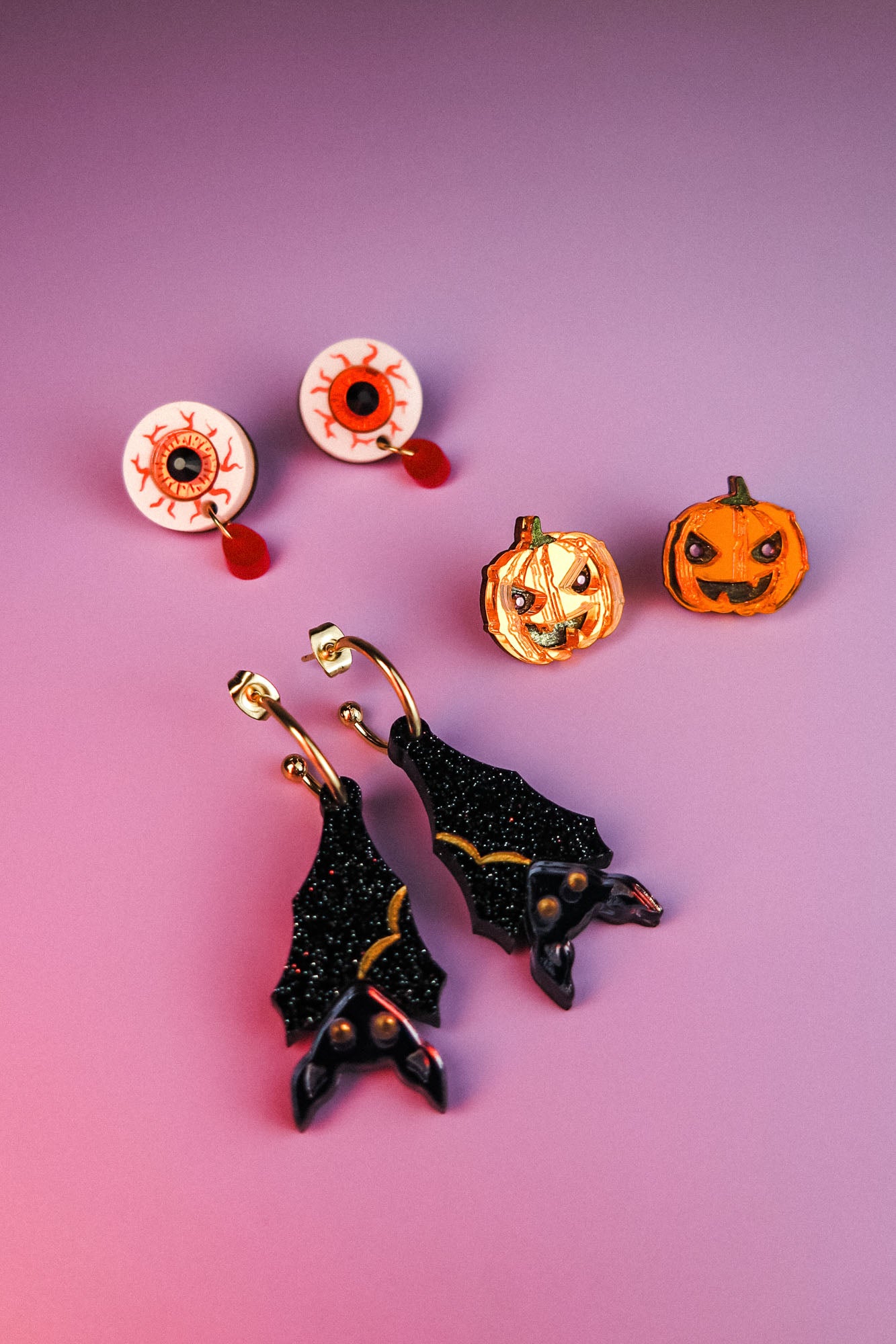 A set of festive Halloween earrings on a purple surface, including hand-painted earrings shaped like eyeballs, jack-o-lantern pumpkins, and sparkly black bats with gold accents—all allergy-free earrings for comfortable wear.
