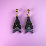 A pair of black hand-painted bat-shaped earrings with yellow accents and orange eyes on gold hooks, displayed on a pastel purple background. These allergy-free earrings add a whimsical touch to any outfit.