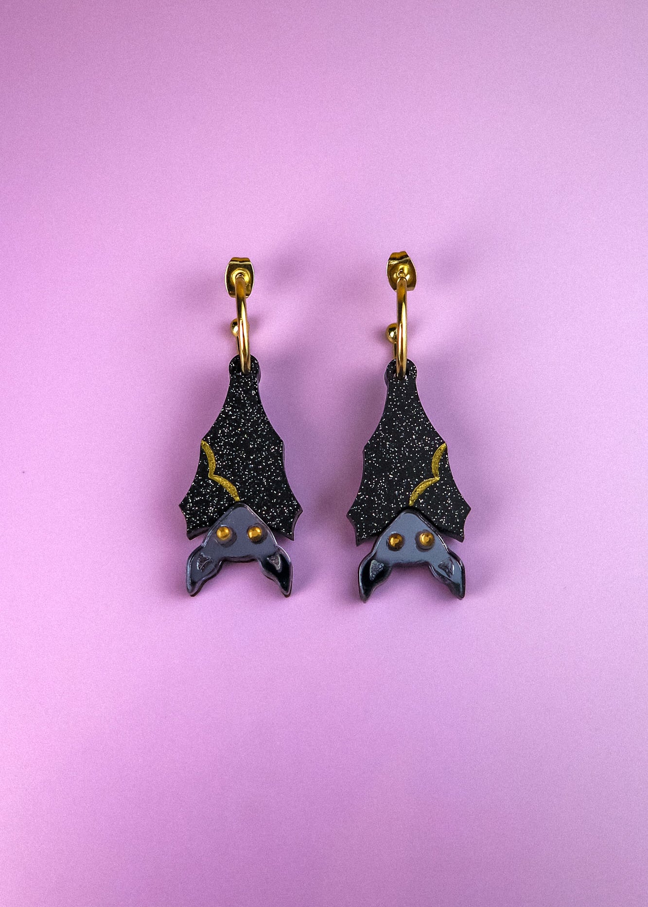 A pair of black hand-painted bat-shaped earrings with yellow accents and orange eyes on gold hooks, displayed on a pastel purple background. These allergy-free earrings add a whimsical touch to any outfit.