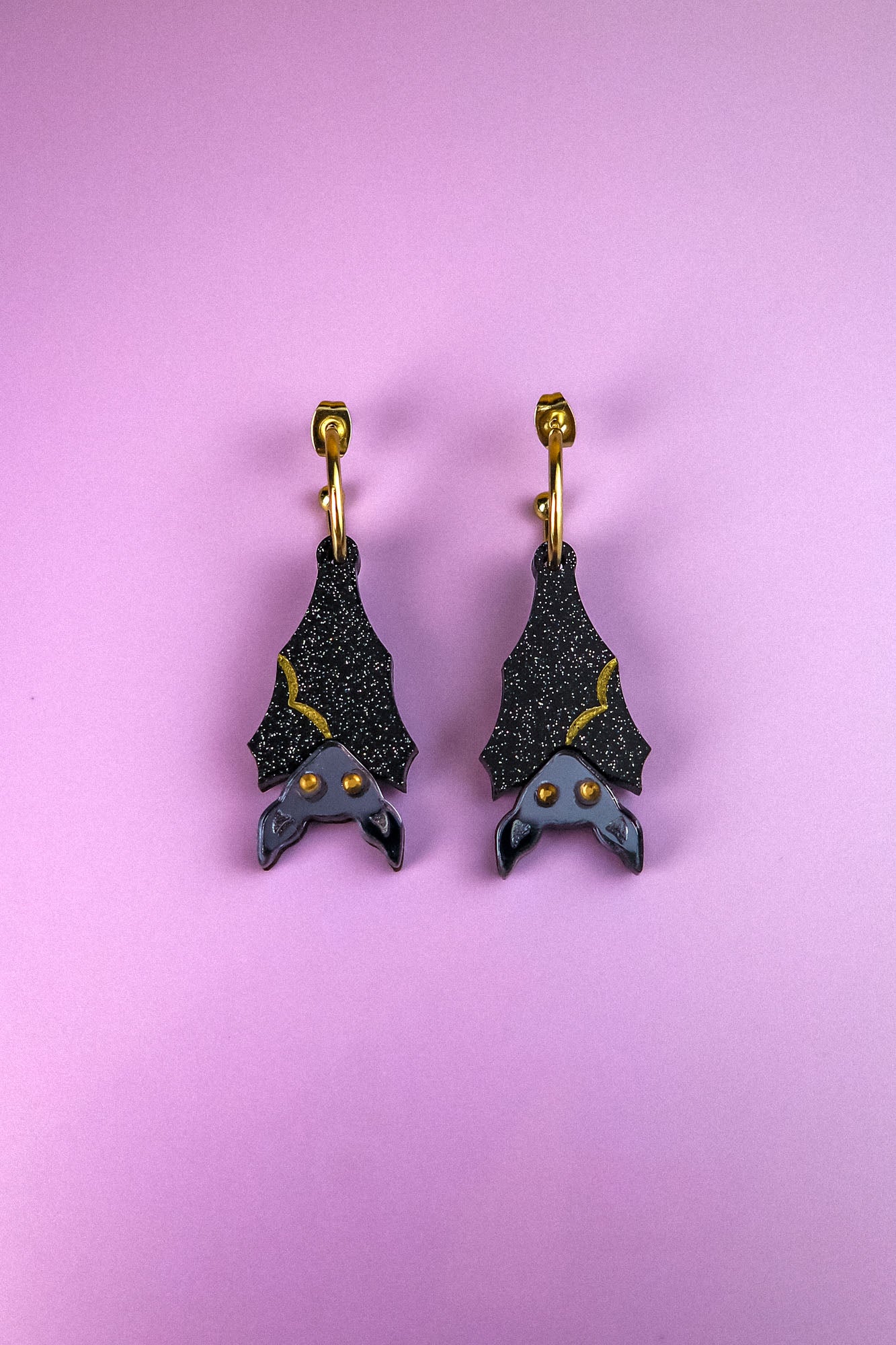 A pair of black hand-painted bat-shaped earrings with yellow accents and orange eyes on gold hooks, displayed on a pastel purple background. These allergy-free earrings add a whimsical touch to any outfit.